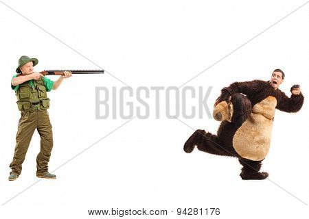 Full length portrait of a mature hunter aiming a rifle towards a man in a bear costume isolated on white background