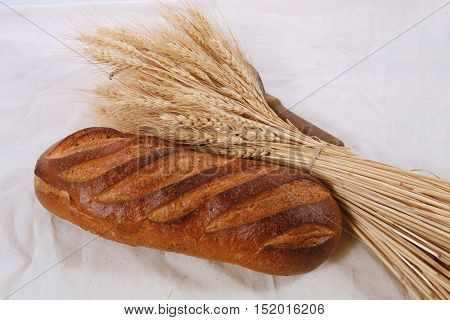 bunch of mown wheat ears with vintage handmade reaper hook sickle and rye dark white french fresh bread loaf on light linen tablecloth