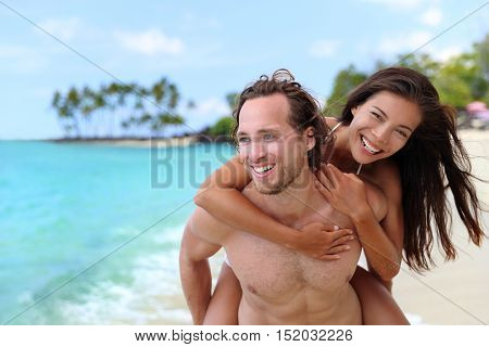 Attractive happy couple laughing having fun piggybacking on tropical beach. Caucasian man carrying Asian girlfriend having fun laughing on travel vacation. Healthy happy interracial people.