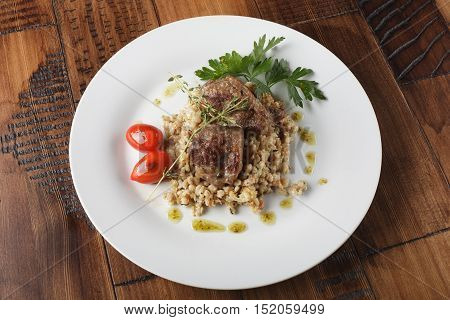 Veal (beef) cheeks with pearl barley porridge and greens in a white plate. Wooden background.