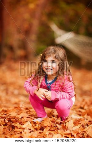 Nice little girl playing in autumn park, sweet kid sitting on the dry brown tree leaves, enjoying beauty of the fall