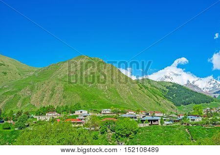 The houses of Stepantsminda resort located at the green mountain foot and the Gergeti Trinity Church next to the snowy and cloudy Kazbek Mount Kazbegi Georgia.
