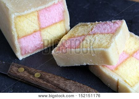 Freshly Cut Battenberg Cake On Dark Slate Background With Knife. Pink And Yellow Sponge Covered In M