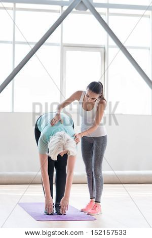 It is more difficult than it seems to be. Active enthusiastic slim woman standing near her coach and bending forwards while doing fitness exercises