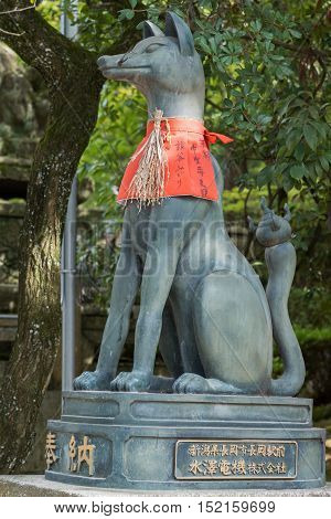 Kyoto Japan - September 17 2016: Fushimi Inari Taisha Shinto Shrine. Kitsune the brown fox messenger and server of God Inari has rice strung around its neck and sports a red apron. Against green tree foliage.