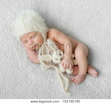bare sleeping baby in hat with toy on white soft blanket with hand under his head
