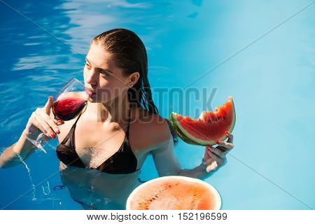 young sexy woman or girl with pretty face and wet hair swimming in pool with blue water eating red watermelon and drinking wine from glass sunny summer day outdoor