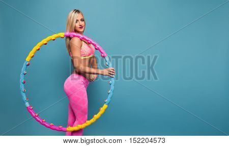 Portrait of adorable blonde woman in pink sportswear holding a hoop while posing on blue background