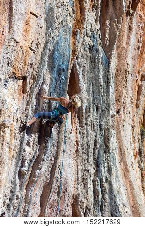 Blond Female Climber ascending Vertical Orange Rocky Wall unusual shape of Mountain Range on background Outdoor Sunshine