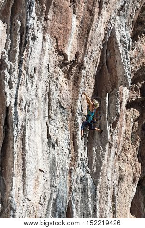 Blond Female Climber ascending Vertical Orange Rocky Wall unusual shape of Mountain Range on background Outdoor Sunshine