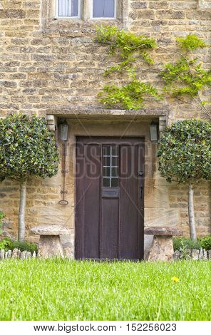 Exterior brown wooden doors of traditional English cottage with topiary bay leaf trees staddle stones grass lawn in front