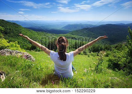 Happy young female traveller sitting at green grass glade at mountain landscape background in sunny spring day under blue cloudy sky with her hands up