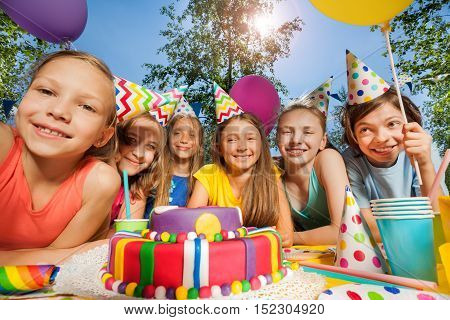 Six happy kids in party hats, standing in a row around birthday cake at the outdoor party