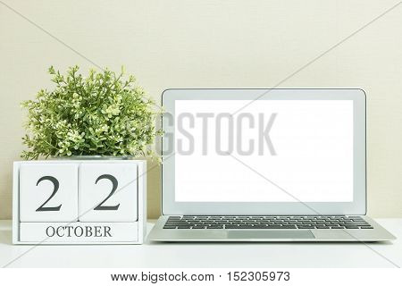 White wooden calendar with black 22 october word with white blank space at center of computer notebook on white wood desk and cream wallpaper textured background selective focus at the calendar