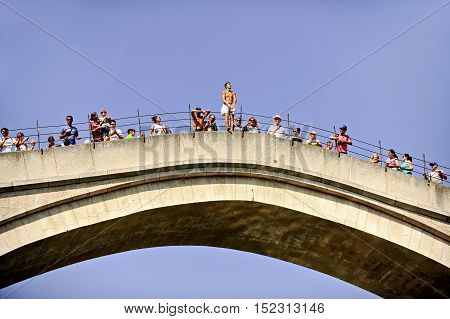 MOSTAR BOSNIA AND HERZEGOVINA - AUGUST 29: Tourists watch a high diver jumping from the old bridge of Mostar into Neretva river on August 29 2015 in Mostar.
