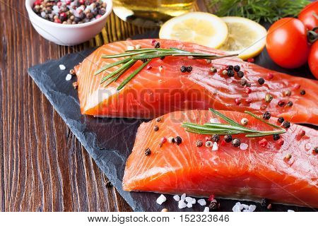 Raw salmon fillet and ingredients for cooking on a slade board and brown wooden background.