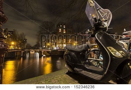 A nightscape in the Amsterdam city center, showing canals lit by streetlights, apartments and scooter parked on the pavement lining the canal.