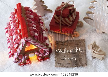 Label With German Text Weihnachtsferien Means Christmas Holidays. Gingerbread House On Snow With Christmas Decoration Like Trees And Moose. Sleigh With Christmas Gifts Or Presents.
