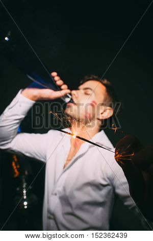 Young attractive man in white shirt drinking from bottle and holding sparkler on the foreground. Adult with red lipstick on his cheek at after party.