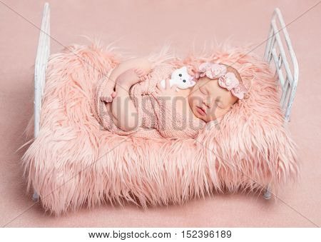 cute sleeping newborn girl with crossed legs and toy cat on little bed with pink fluffy blanket