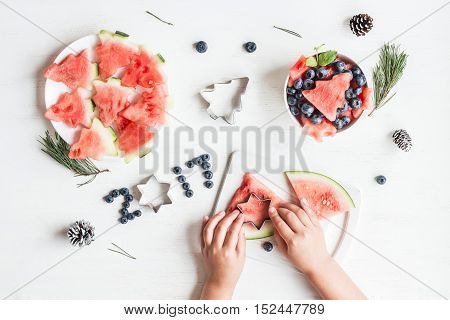 Christmas salad of watermelon and blueberries for kids. Children's hands cooking christmas fruit salad on white table. Top view flat lay