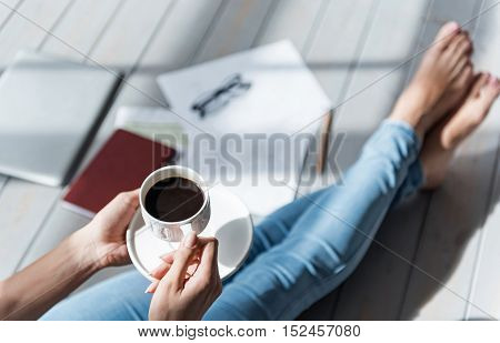 Small pause. Top view of lady sitting barefoot on grey wooden floor and holding full coffee cup.