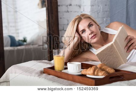 Relaxing moment. Pretty blond lady lying on bed during breakfast time and holding opened book .