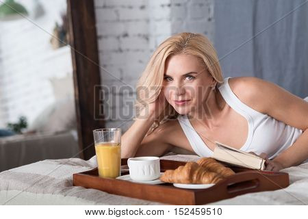Time to eat. Beautiful blond woman lying on bed with closed book and going to have breakfast.