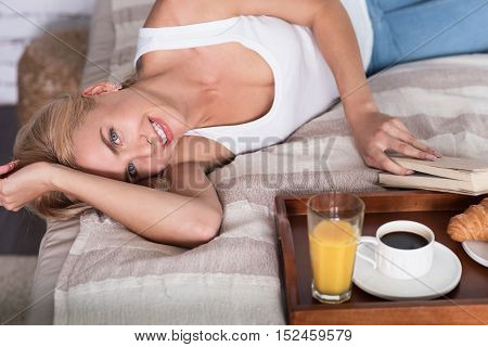 Just happy. Vivacious young blond woman lying on bed near tray with breakfast and holding book.
