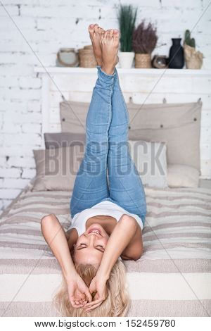 Having fun. Smiling blond lady lying on big bed in leght room and holding legs up.