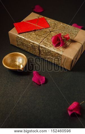 A beautiful gift pack wrapped in a golden paper, tied with golden thread. An elegant Diwali gift background with earthen lamp and flowers.