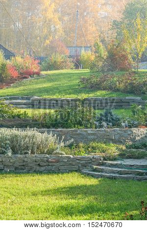 Beautiful autumn landscape with green lawns retaining walls flower beds and a fragment of an alpine slide on the background of yellow leaves of birch trees on a sunny October day
