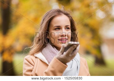 season, technology and people concept - beautiful young woman walking in autumn park and using voice command recorder on smartphone