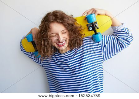 A young redhead woman is standing with yellow colorful skateboard behind her head. Hipster girl laughing with joyful feelings over white wall. Carefree lifestyle