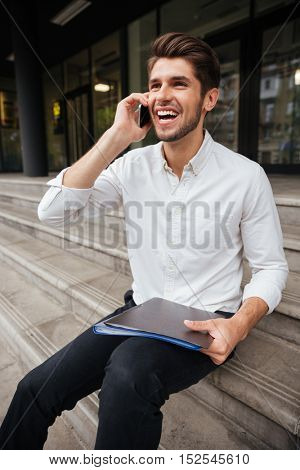 Happy young businessman holding folder with documents and talking on cell phone outdoors