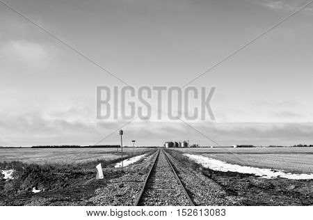 Straight railway tracks dividing harvested fields in late fall with grain bins in the distance in rural black and white landscape in saskatchewan