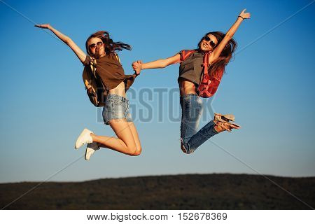 two pretty sexy cute girls or women in jeans and shorts with backpacks outdoor on sunny day jumping on blue sky background