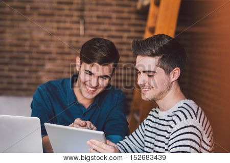Smiling young friends sitting at a table at home using a laptop and digital tablet while working together on their home based business