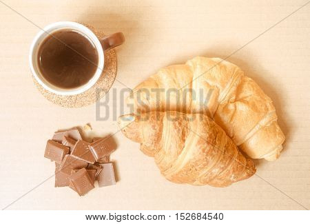 Two baked croissants with a cup of coffee and chocolate bars on white paper's surface.