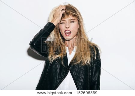 Enjoying freedom. Funky young woman in leather coat holding hand in hair and sticking out tongue while standing against white background