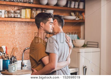 Affectionate young gay couple hugging each other with their eyes closed while standing in their kitchen in the morning