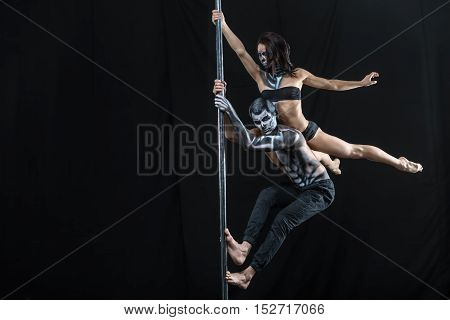 Awesome couple of pole dancers with a horrific body-art in the dark studio. Guy hangs on a pylon and a girl lies on his back and holds the pylon with her right hand. She lies with outstretched legs.