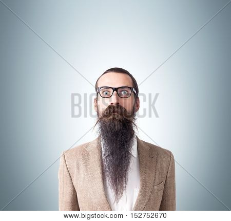 Baffled man wearing glasses and long beard is standing against gray background. Concept of eccentric person. Mock up