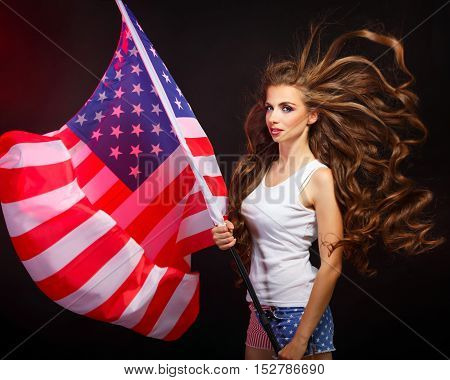 Young attractive girl holding a US flag on the flagpole. She a T-shirt and shorts with a print of the flag. Long curly hair. Portrait of a real patriot. The Star Stripes.