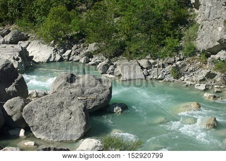 Clear pale blue fast flowing river with large rocks and rapids near the village of Rimon in the Drome valley France. Trees lining the river as background.