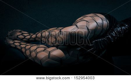 Low key special toned photo of sexy female nude legs in net tights and hands in black leather gloves against dark background horizontal view