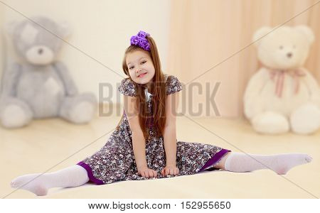 Caucasian little girl with a big purple bow on her head. Girl shows how to do the splits.In the nursery with Teddy bears.