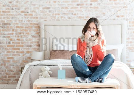 Winter epidemics. Nice moody young woman sitting in a bedroom and wearing a scarf while suffering from cold