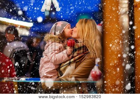Beatiful mother and little daughter eating crystalized sugared apple on German Christmas market. Happy family in winter clothes with lights on background. Family, tradition, holiday concept
