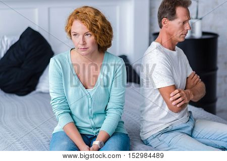 Quarrelsome. Irritated husband and wife sitting back to back on the bed with crossed arms